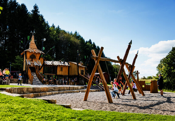 Das Bild zeigt Kinder, die auf de Spielplatz mit Rutschen, Schaukeln und Klettergerüsten spielen.