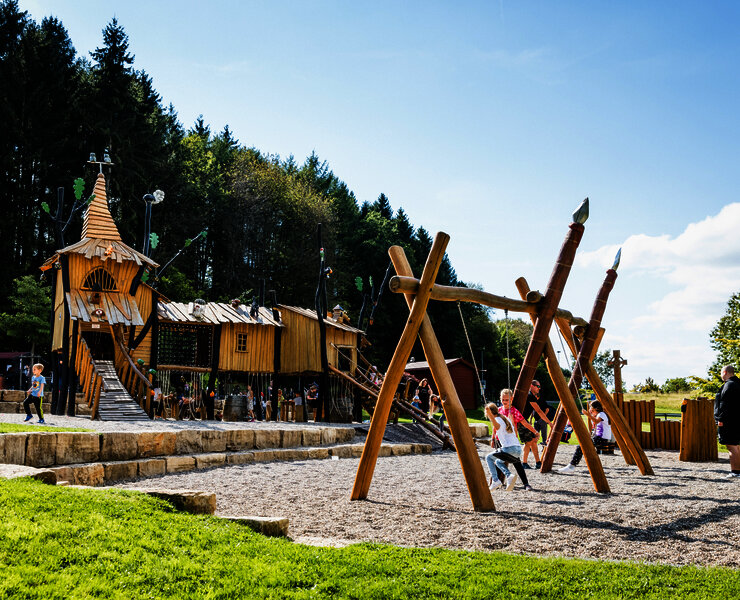Das Bild zeigt Kinder, die auf de Spielplatz mit Rutschen, Schaukeln und Klettergerüsten spielen.
