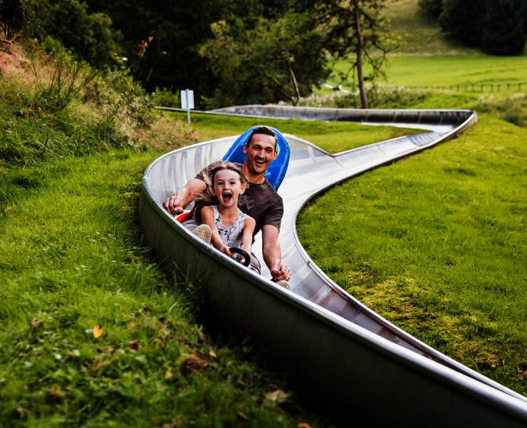 Das Bild zeigt ein Mann und ein Mädchen, die eine Sommerrodelbahn fahren.