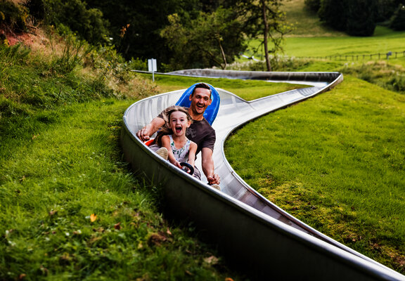 Das Bild zeigt ein Mann und ein Mädchen, die eine Sommerrodelbahn fahren.