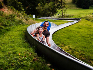 Das Bild zeigt ein Mann und ein Mädchen, die eine Sommerrodelbahn fahren.