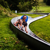 Das Bild zeigt ein Mann und ein Mädchen, die eine Sommerrodelbahn fahren.