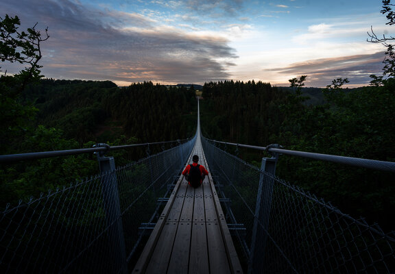 Das Bild zeigt eine Person die mittig auf der Hängeseilbrücke Geierlay sitzt. Rund um die Brücke befindet sich tiefgrünes Waldgebiet.