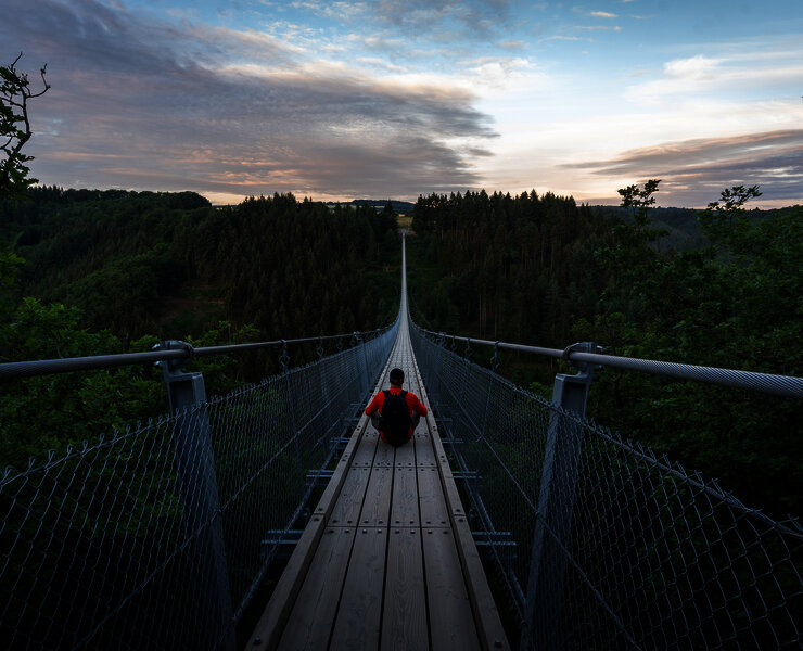 Das Bild zeigt eine Person die mittig auf der Hängeseilbrücke Geierlay sitzt. Rund um die Brücke befindet sich tiefgrünes Waldgebiet.