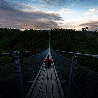Das Bild zeigt eine Person die mittig auf der Hängeseilbrücke Geierlay sitzt. Rund um die Brücke befindet sich tiefgrünes Waldgebiet.