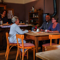 Das Bild zeigt die Gaststube des Hunsrücker Holzmuseums. Drei Personen sitzen am Tisch und trinken Kaffee. Eine Person steht hinter der Theke, auf der auch Kuchen steht.