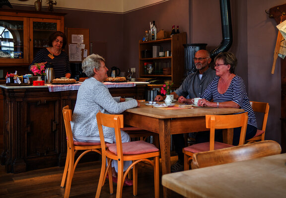 Das Bild zeigt die Gaststube des Hunsrücker Holzmuseums. Drei Personen sitzen am Tisch und trinken Kaffee. Eine Person steht hinter der Theke, auf der auch Kuchen steht.