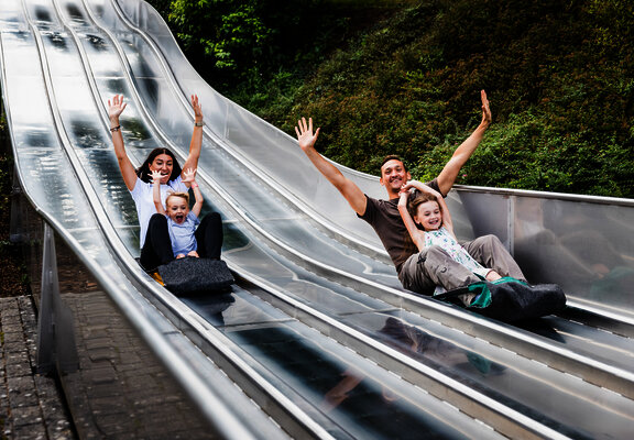 Das Bild zeigt zwei Erwachsene und zwei Kinder, die auf einer Teppichrutsche beim Freizeitzentrum Peterberg rutschen.