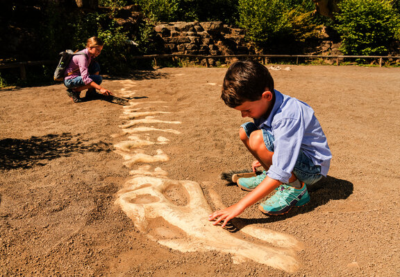 Das Bild zeigt zwei Kinder im Dinopark Teufelsschlucht, die auf einer Sandfläche ein Dinoskelett ausgraben.