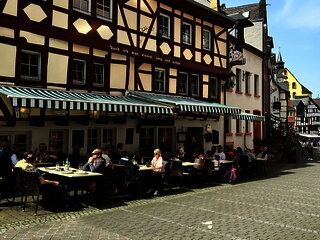 Ansicht auf das Restaurant Alt Bernkastel mit Fachwerkfassade und Außenbestuhlung, wo Gäste in der Sonne sitzen.